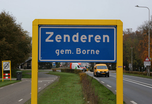 Bord van Zenderen gemeente Borne met gele ophanging. Bord staat naast een weg waar auto's aan komen rijden