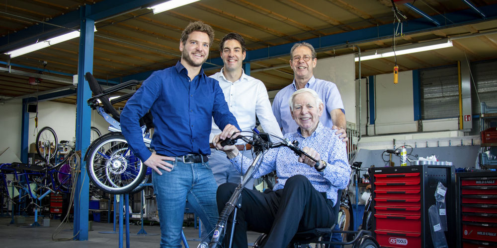 Drie mensen staan om een man die zit op een driewieler in een fietsenmakerswinkel