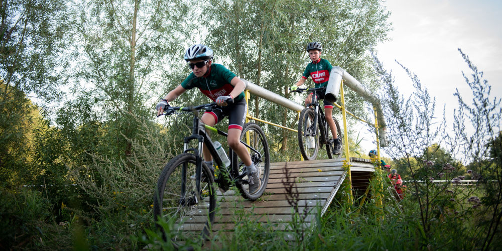 Twee fietsers fietsen over houten brug tussen hoog gras en bomen